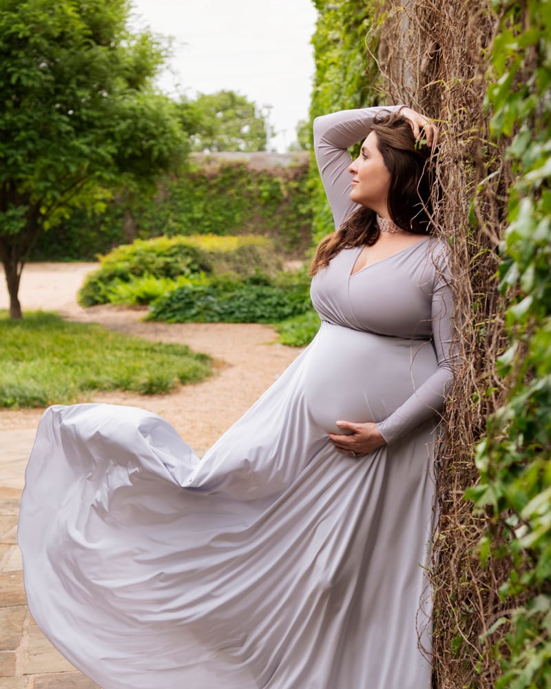 Maternity Photographer, woman in flowing dress leans agains ivy covered wall in a park