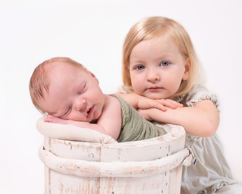 Newborn Photography, a toddler older sister leans behind her new baby sibling who sleeps softly in a wooden bucket with linens