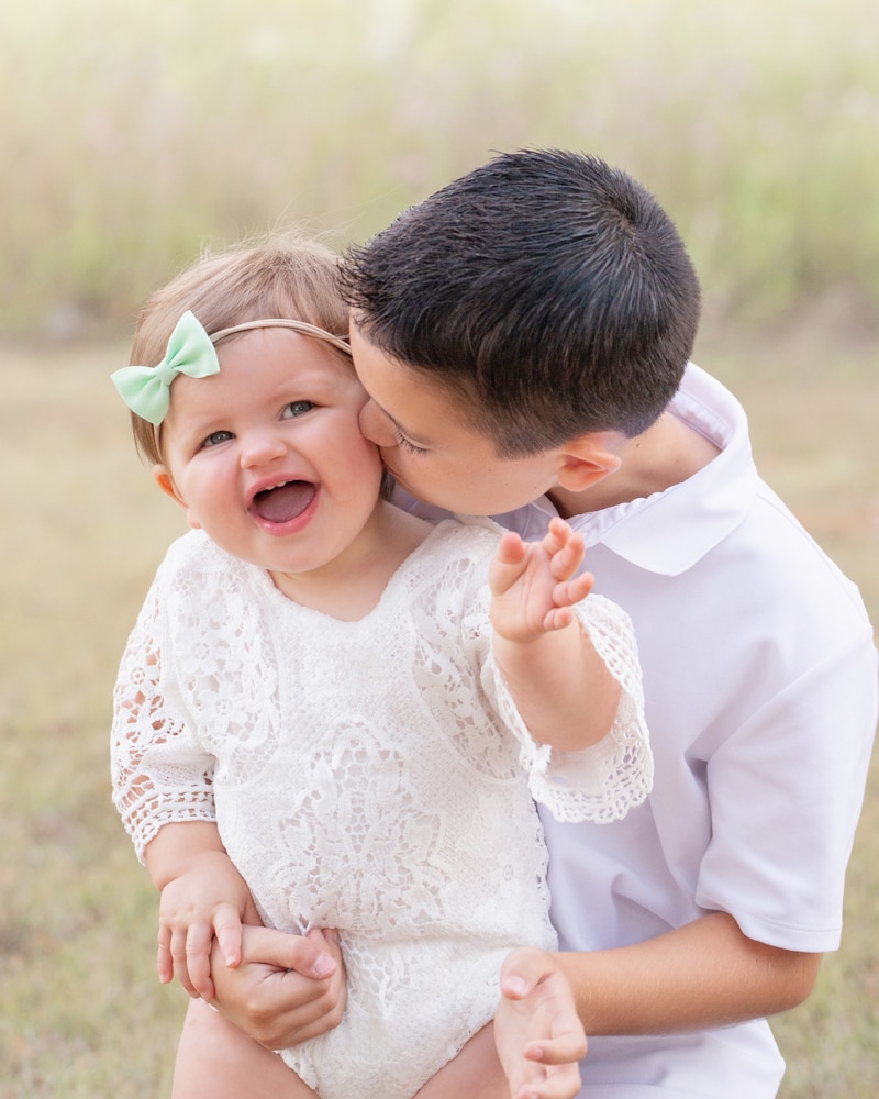 milestone photography, older brother kisses his baby sister on the cheek outside