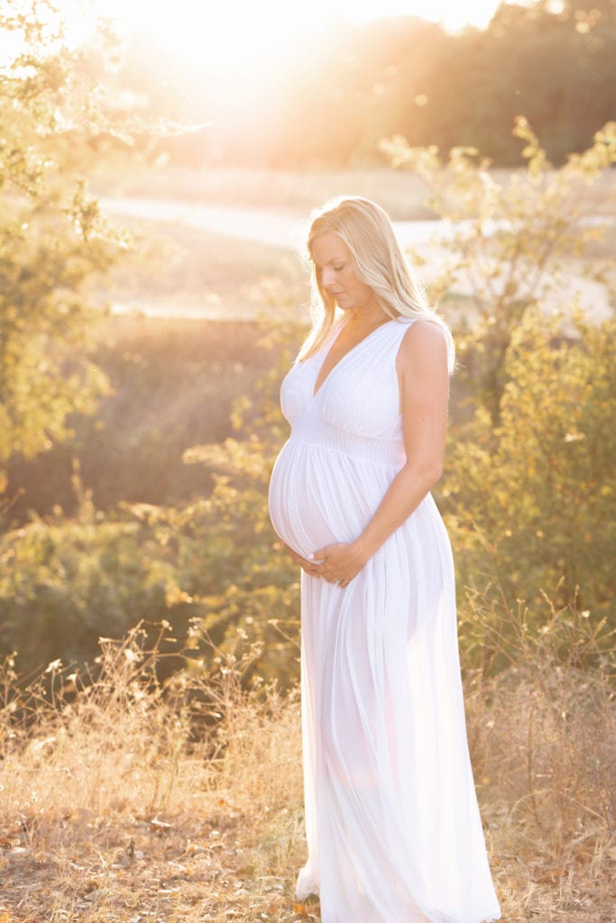 Maternity photography, A woman stands in the grass near trees, she holds her belly, she is pregnant