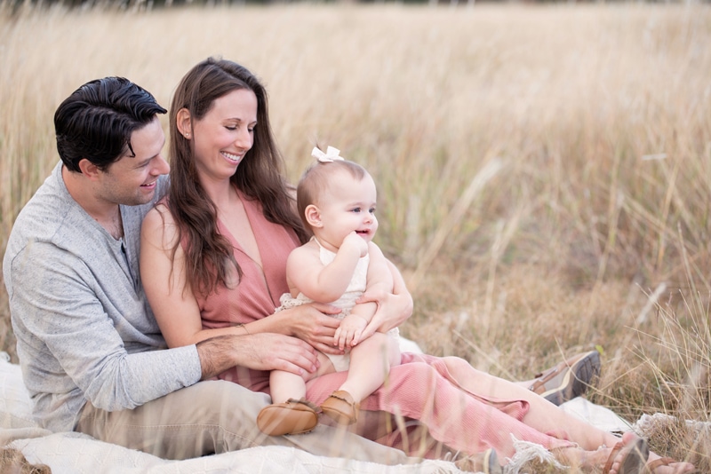 a family of 3, a man and woman hold onto their baby girl as they all sit in a field outside