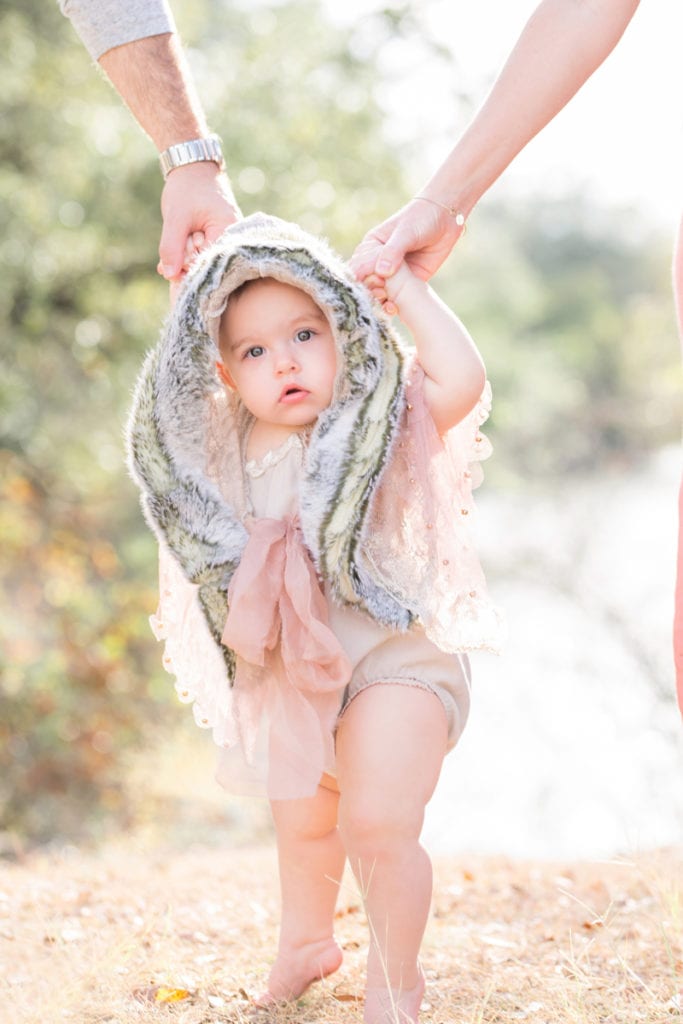 A baby holds her parents hands outdoors, she is wrapped in a furry hood