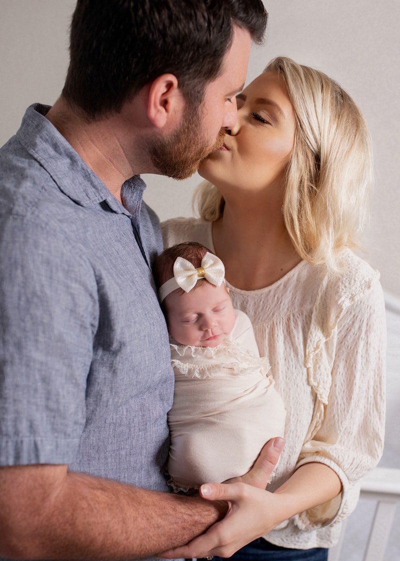 Newborn Photography, a husband and wife kiss as they hold their swaddled baby girl