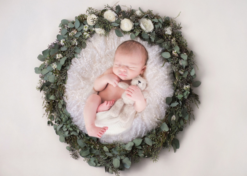 Newborn Photography, a little baby holds a teddy bear and lays in a cozy bed with a wreath around it